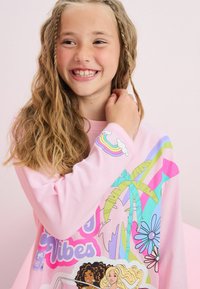 Ljusrosa sweatshirt med färgglada grafiska teman, inklusive palmträd och blommor, med fet text som säger "Good Vibes". Mjuk material.