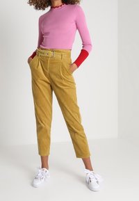 Eng anliegendes rosa Langarmoberteil mit roten Bündchen, kombiniert mit hoch taillierter gelber Cordhosen und weißen Sneakers mit Schnürsenkeln.