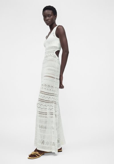 Ένα ψηλό άτομο στέκεται φορώντας ένα λευκό, αμάνικο, πλεκτό μάξι φόρεμα με πλαϊνές ανοίγματα και μπεζ επίπεδα σανδάλια, μπροστά σε μια απλή φόντο.