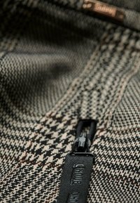 Gros plan sur un tissu pied-de-poule marron et noir avec un tirette de fermeture éclair noire gravée du mot "LIU JO".