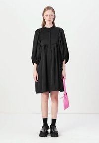 Vestido preto com mangas bufantes, decote redondo, corte descontraído e comprimento até ao joelho. Estilizado com uma bolsa rosa e sapatos de plataforma.