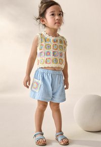 Stricktop ohne Ärmel mit bunten Granny-Quadrat-Mustern; hellblaue Shorts mit elastischem Bund und aufgesetzter Tasche; blaue Sandalen.