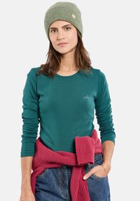 Teal lange mouwen t-shirt met ronde hals en een klein logo, gecombineerd met een roze gebreide trui om de taille geknoopt en een groene geribbelde beanie.