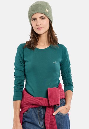 T-shirt long à manches teal avec un col rond et un petit logo, associé à un pull en tricot rose noué à la taille et un bonnet côtelé vert.