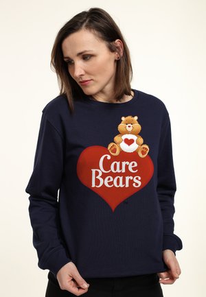 Donna che indossa una felpa blu navy con un cuore rosso e un personaggio dei Care Bears che tiene un cuore sul petto.