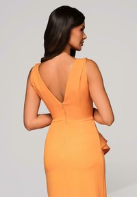 Vrouw met lang donker haar, gekleed in een mouwloze oranje jurk met een diepe V-hals aan de achterkant en een zijruffle, staande tegen een effen achtergrond.