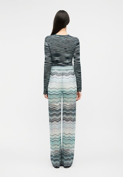 Žena s dlhými tmavými vlasmi, oblečená v pruhovanom top s dlhým rukávom a vysokým pásom širokými nohavicami so vzorom cik-cak, otočená chrbtom ku kamere.