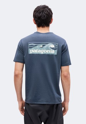 Uomo visto da dietro che indossa una maglietta Patagonia blu con un motivo a onda e testo nella parte superiore della schiena.