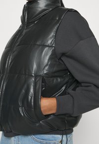 Svart vadderad väst gjord av glansigt material, med hög krage, framdragkedja, sidofickor och elastiska muddar, kombinerad med en mörk sweatshirt.