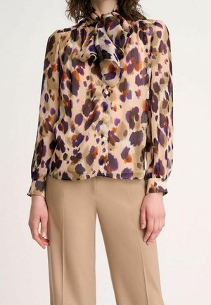 Kobieta ubrana w beżową bluzkę z fioletowymi, brązowymi i pomarańczowymi abstrakcyjnymi plamami, ozdobioną dużą kokardą przy dekolcie, w połączeniu z beżowymi spodniami.