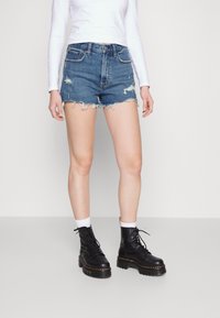 Pantalones cortos de mezclilla azules con dobladillo deshilachado y detalles desgastados, llevados con botas negras con cordones y calcetines blancos por encima del tobillo.