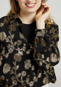 Blouse noire avec un motif floral métallique doré. Manches longues avec des poignets élastiques, design avec col, tissu texturé léger.