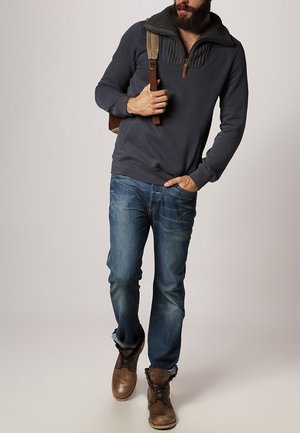 Homme barbu portant un pull sombre avec col zippé, jean bleu, bottes marron à lacets, portant un sac à dos beige sur une épaule.