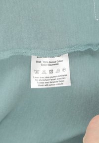Tessuto di cotone in un morbido color teal, con un'etichetta di cura che dettaglia le istruzioni di lavaggio in più lingue. La cucitura è rifinita con precisione.
