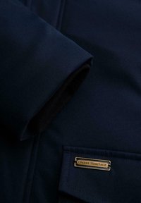 Marineblauwe jas van gladde stof, met een manchet met een zwarte voering en een metalen label waarop "OMBRE HERITAGE" op de zak staat.