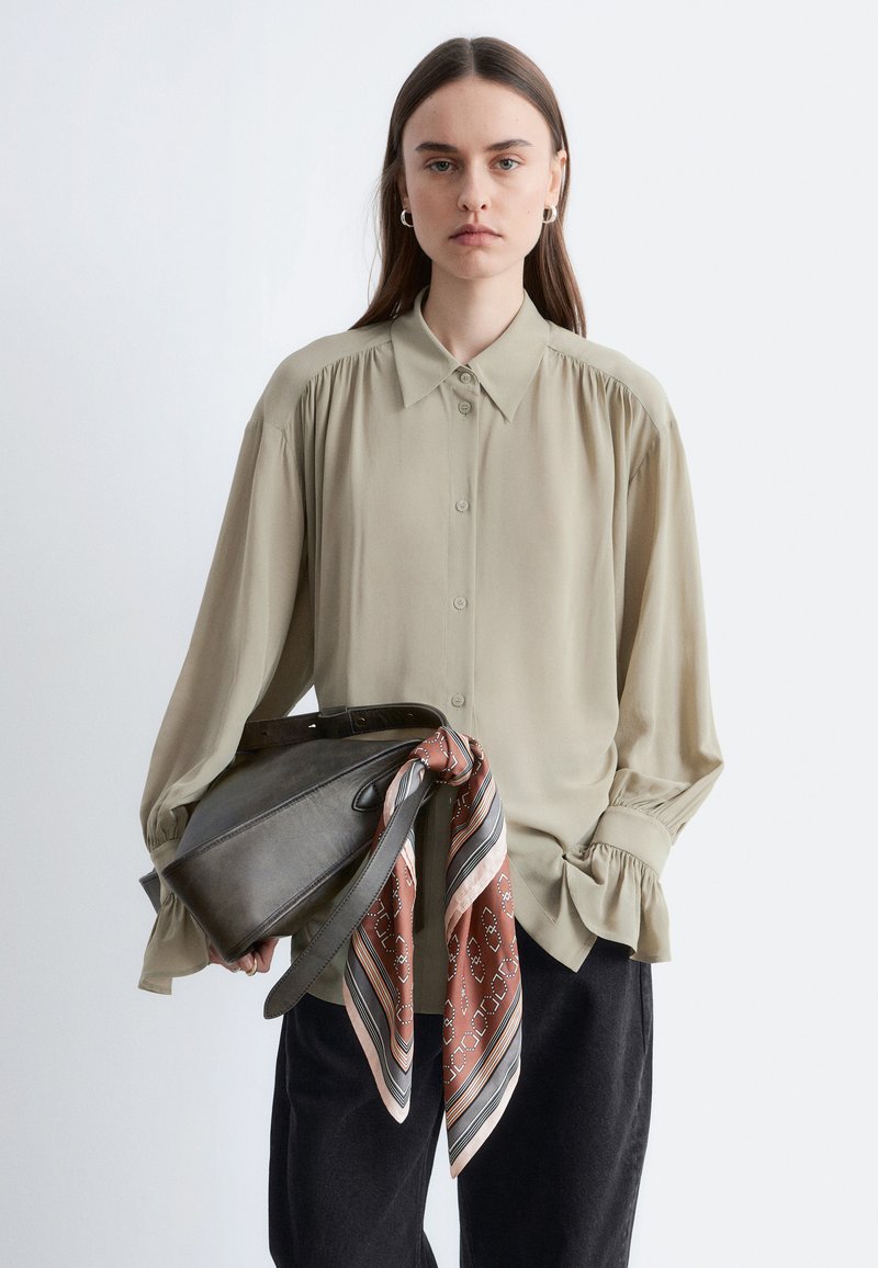 Femme aux cheveux bruns raides, portant une chemise beige ample boutonnée et un pantalon noir, tenant un sac à main en cuir gris foncé avec une écharpe marron à motifs.