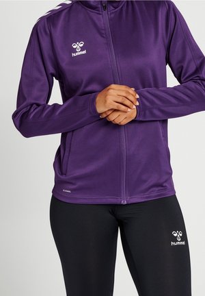 Kvinde iført en lilla sportjakke med lynlås og sorte leggings, begge med hvide hummel-logoer, stående med hænderne sammenknyttede.