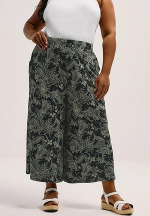 Frau trägt schwarze weite Hosen mit weißem floralen Paisley-Muster, weißes ärmelloses Oberteil und weiße Keilsandalen, steht mit Hand in der Hüfte.