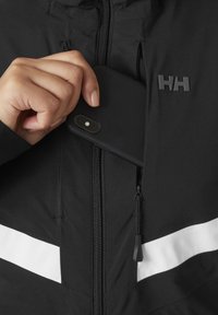 Schwarze Jacke mit einem Reißverschluss vorne, ausgestattet mit einem weißen Streifen und einem geprägten Logo. Eine Hand legt ein schwarzes Handy in eine mit Reißverschluss verschlossene Tasche.