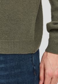 Maglione lavorato a maglia verde oliva con orli e dettagli a costine, abbinato a jeans in denim blu scuro. La texture del maglione è morbida e leggermente pelosa.
