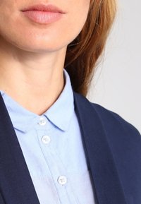 Granatowa marynarka na jasnoniebieskiej koszuli z kołnierzykiem, ze szczegółami guzików. Marynarka ma gładką teksturę i zorganizowany krój.