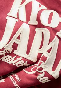 Gros plan sur un tissu rouge avec de grandes lettres blanches en relief formant les mots « KYOTO JAPAN » et un texte en cursive plus petit « Easter » sur un vêtement.