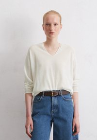 Cremfarbener V-Ausschnitt-Pullover mit langen Ärmeln, kombiniert mit hochtaillierten blauen Jeans und einem braunen Ledergürtel mit goldener Schnalle.