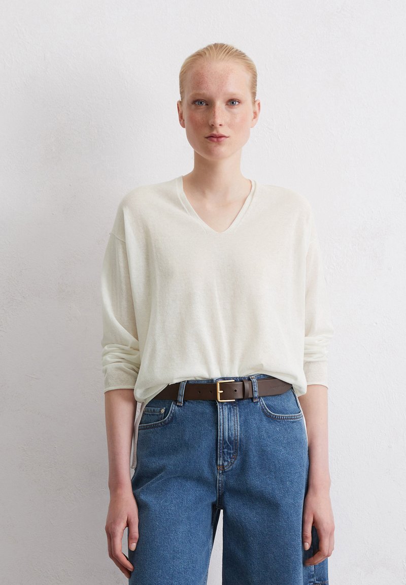 Cremfarbener V-Ausschnitt-Pullover mit langen Ärmeln, kombiniert mit hochtaillierten blauen Jeans und einem braunen Ledergürtel mit goldener Schnalle.