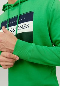 Osoba upravující rukáv jasně zelené mikiny s kapucí s černým, bílým a tmavozeleným obdélníkovým logem a textem "JACK & JONES" na hrudi.