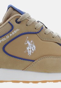 Ljusbruna sneakers med ovandel i mesh och syntetmaterial, blå detaljer och en vit polo-spelare logotyp på sidan. Har beige skosnören och sula.