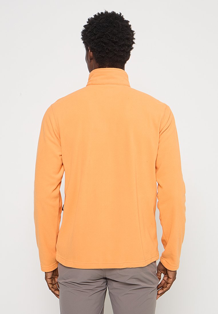 Mężczyzna tyłem, ubrany w jasnopomarańczową polarową kurtkę i jasnoszare spodnie na jednolitym białym tle.