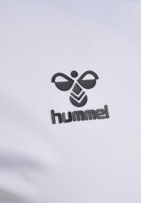 Vitt tyg med en texturerad yta som har en svart Hummel-logotyp, inklusive stiliserade vingar och tre nedåtgående chevroner.
