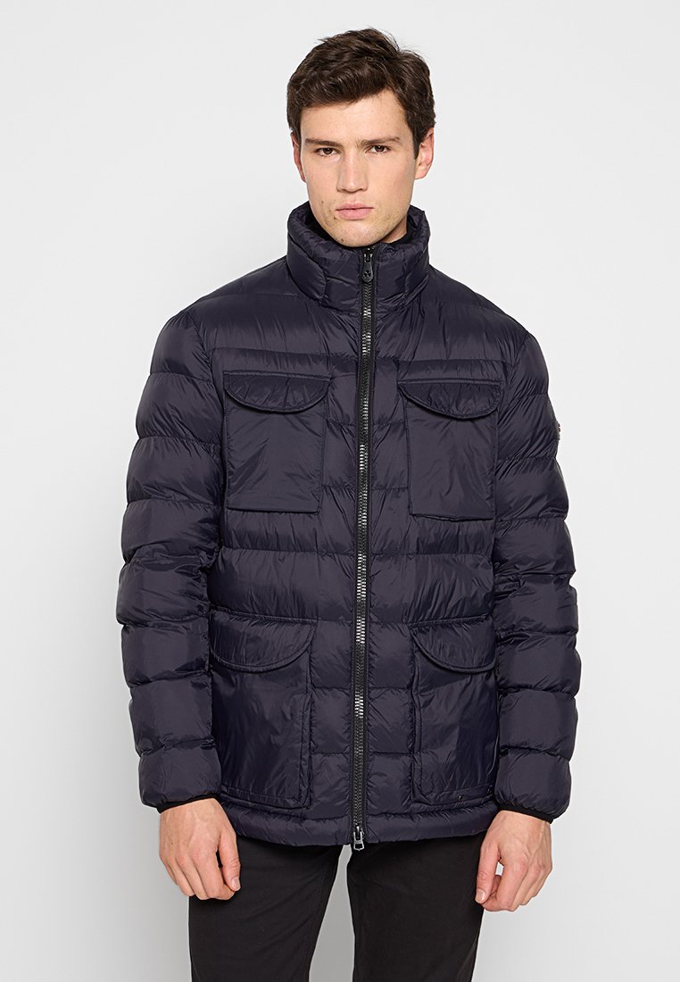 Man in een zwarte gewatteerde jas met een rits aan de voorkant en vier klepzakken, staande tegen een effen witte achtergrond.