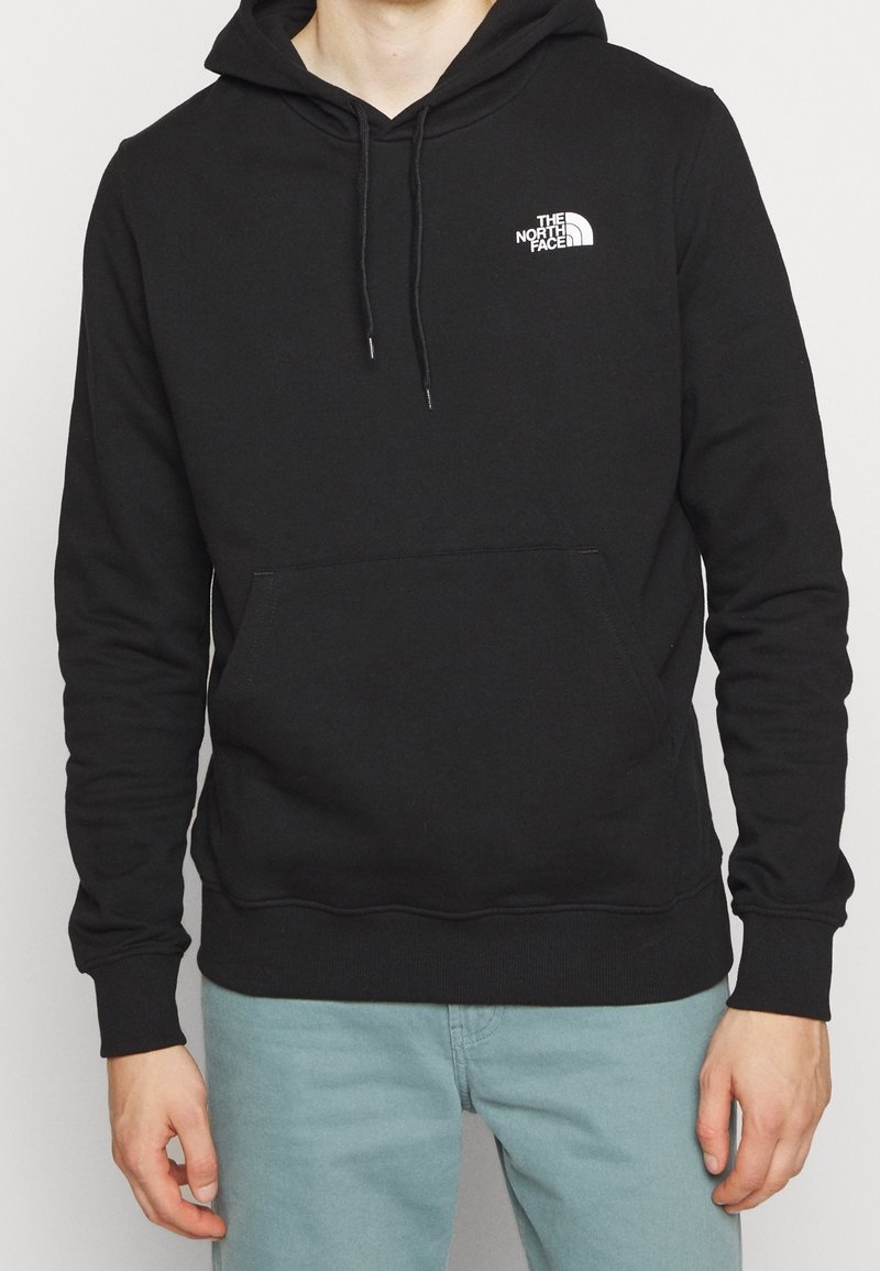 Personne portant un sweat à capuche noir The North Face avec poche frontale et capuche, associée à un pantalon bleu clair, debout devant un fond uni.