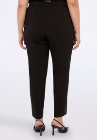 Kvinna som bär svarta skräddarsydda byxor och svarta högklackade slingbackskor, stående mot en enfärgad ljus bakgrund.