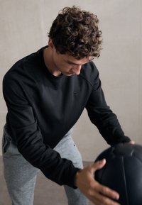 Sweatshirt noir à manches longues et col rond, associé à un pantalon gris clair, tandis qu'une personne tient un ballon de médecine noir.