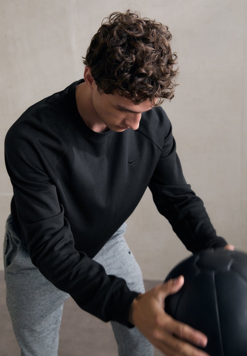 Sweatshirt noir à manches longues et col rond, associé à un pantalon gris clair, tandis qu'une personne tient un ballon de médecine noir.