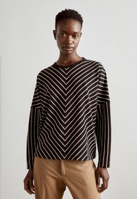 Black and white striped long-sleeve sweater featuring a chevron pattern, round neckline, and relaxed fit. Worn with beige trousers.