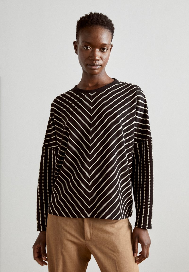 Black and white striped long-sleeve sweater featuring a chevron pattern, round neckline, and relaxed fit. Worn with beige trousers.