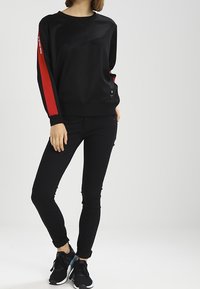 Sudadera negra con rayas laterales rojas, cuello redondo y puños acanalados. Combinada con jeans ajustados negros y zapatillas deportivas negras.