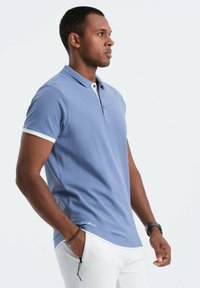 Ljusblå pikétröja i texturerat tyg, med vita detaljer på kragen och ärmarna, samt en liten logotyp vid fållen.