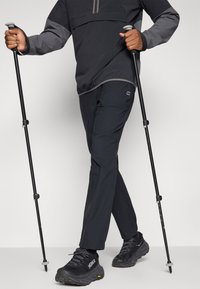 Schwarze Wandershosen und eine Jacke mit grauen Akzenten, kombiniert mit schwarzen Wanderschuhen und Trekkingstöcken. Leichte, funktionale Outdoor-Bekleidung.