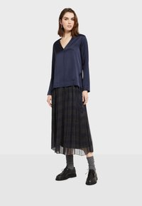 Navy plavi satenski top s dugim rukavima uparen s naboranom crnom kariranom suknjom. Model nosi sive čarape i crne cipele na vezanje.