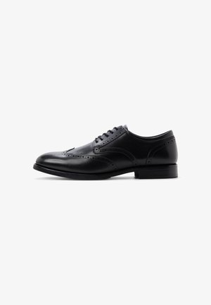 Zwarte leren herenschoenen met brogue-details, gesloten vetersluiting en een lage gestapelde hak op een witte achtergrond.