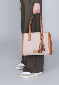 Tan ja valkoinen herringbone-kuviollinen kassi, jossa on nahkaosat ja tupsuyksityiskohta, varustettuna kahdella tukevalla kahvalla ja suorakulmaisella muodolla.