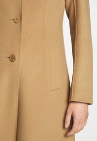 Manteau couleur camel avec deux boutons visibles et une poche latérale, porté par une personne avec la main droite détendue le long du côté.