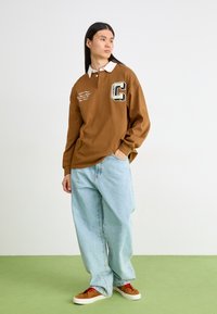Camisa de polo de manga larga marrón con un collar blanco y un gran parche de la letra "C". Combinada con vaqueros sueltos de color azul claro y zapatillas marrones.