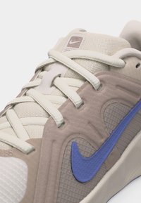 Μπεζ και καμηλό αθλητικό παπούτσι με κορδόνια σε κρεμ απόχρωση, ύφασμα από δίχτυ και μπλε λογότυπο Nike στη πλευρά.