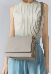 Sac à bandoulière en cuir beige avec une surface texturée, doté d'un rabat et d'un quincaillerie dorée, associé à un fond bleu clair.
