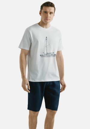 Mężczyzna w białej koszulce z szkicem żaglówki i ciemnoniebieskich szortach, stojący na jasnym, jednolitym tle.